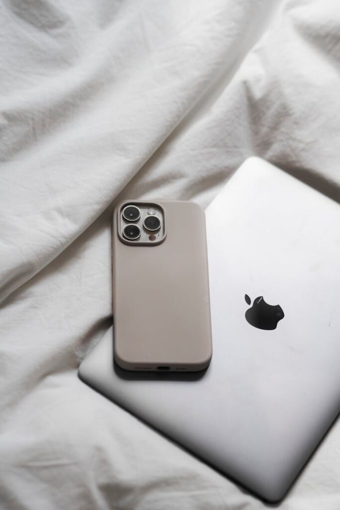 An iPhone and MacBook resting on white bed sheets, showcasing modern technology in a minimalist setting.